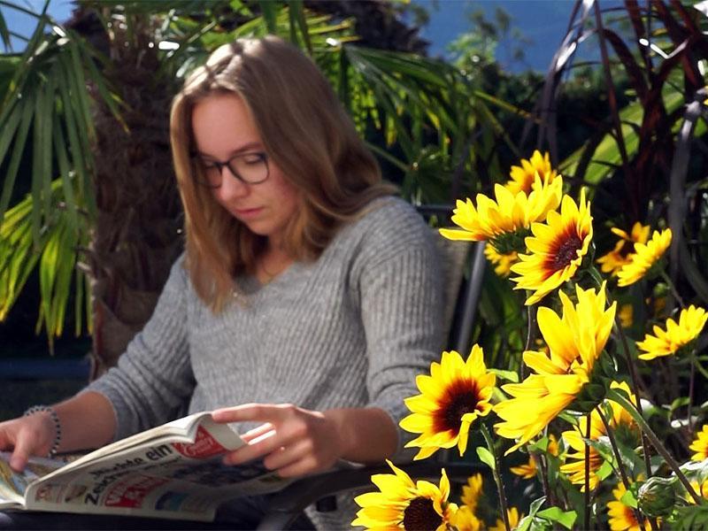 Frau liest Zeitung neben Sonnenprinzessin (Sonnenblumen) auf Balkon